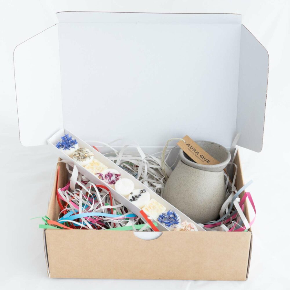 Image of scented gift hamper with open box containing stoneware oil burner and wax melts nestled in recyclable packing paper ribbons